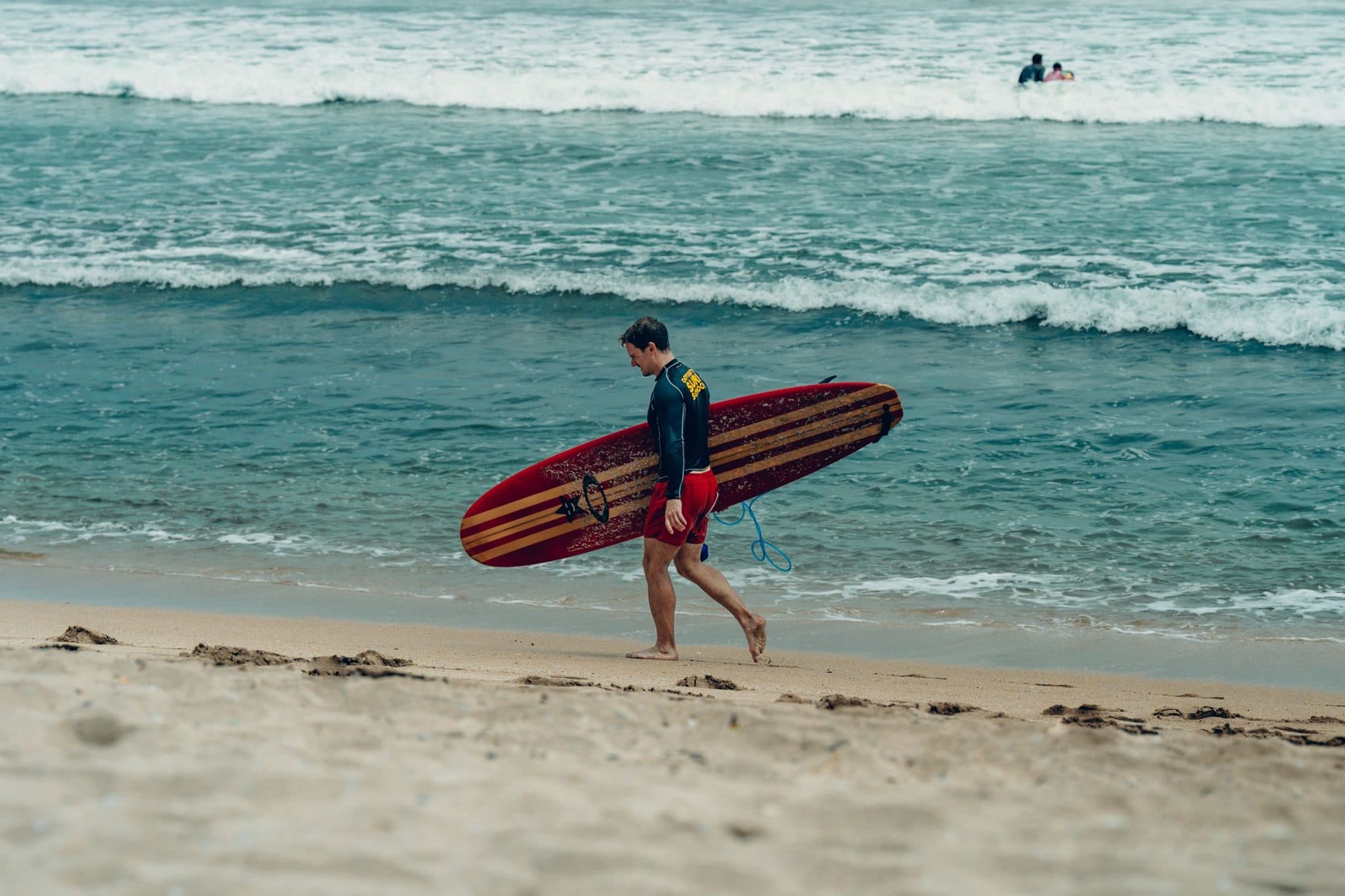 A surfer carrying a red board along the shoreline